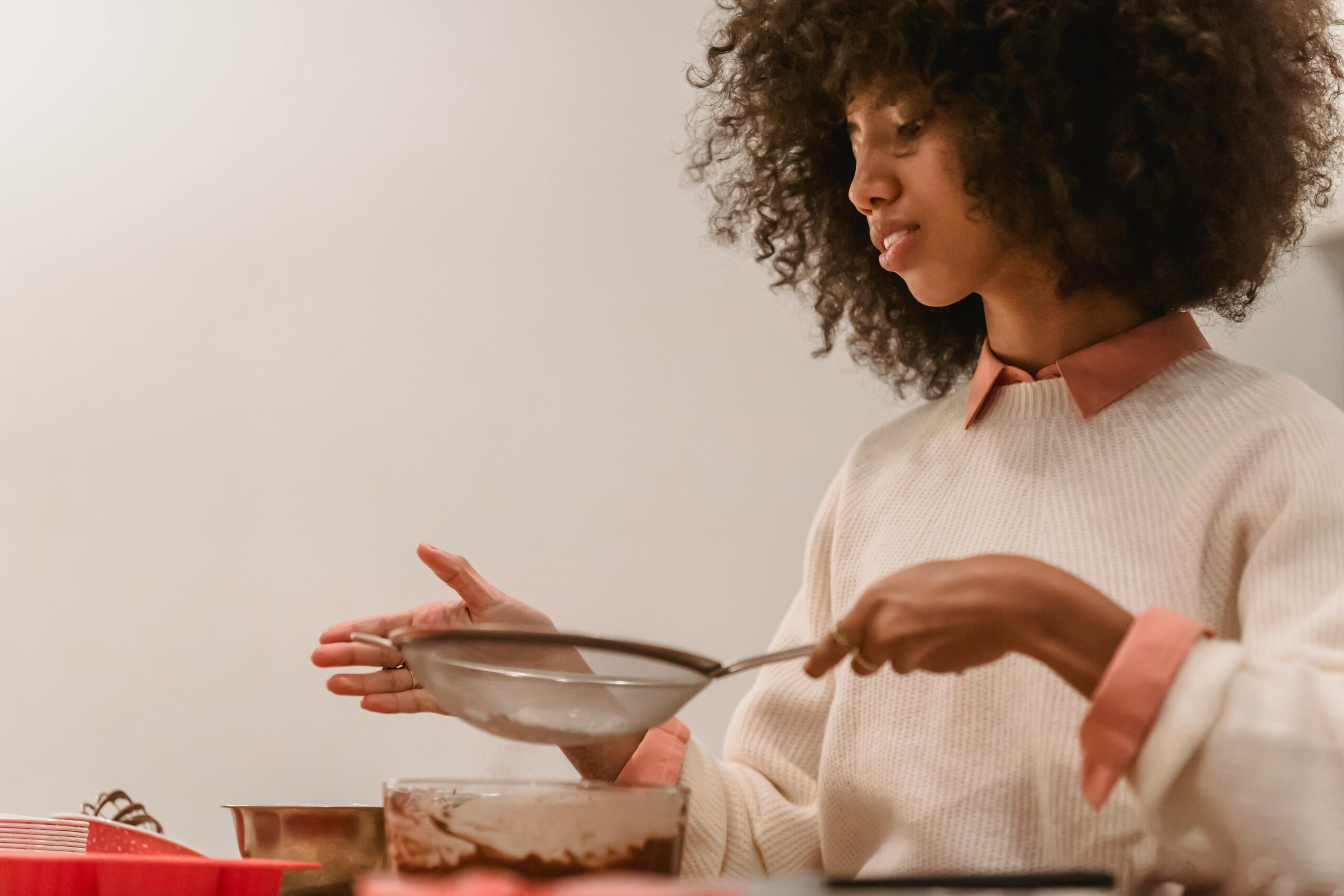 Side view of African American female cook with sieve pouring flour into bowl with chocolate batter while cooking in kitchen